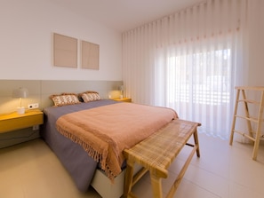 Ein Schlafzimmer mit einem Bett, Nachttischen, einer Bank, einer Leiter und einem Fenster mit durchsichtigen Vorhängen.