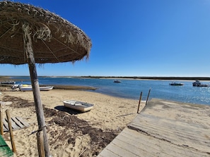 A beach with a wooden deck, a thatched umbrella, and several boats on the water. - Apartment with pool - 4 guests (Tavira)