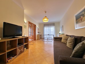 A living room with a flat-screen TV, wooden shelving, a sofa, a dining table with chairs, and a pendant light. - Bright apartment near beach - couples/families (Tavira)