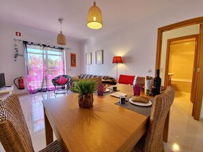 Ein modernes Wohnzimmer mit einem Holzespeisetisch, Korbstühlen, einer Couch und einem großen Fenster mit transparenten Vorhängen.