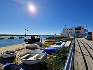 A coastal scene with boats docked at a pier, a sandy beach, and a building with a flag. - Apartment with pool and garden - 4 guests (Tavira)