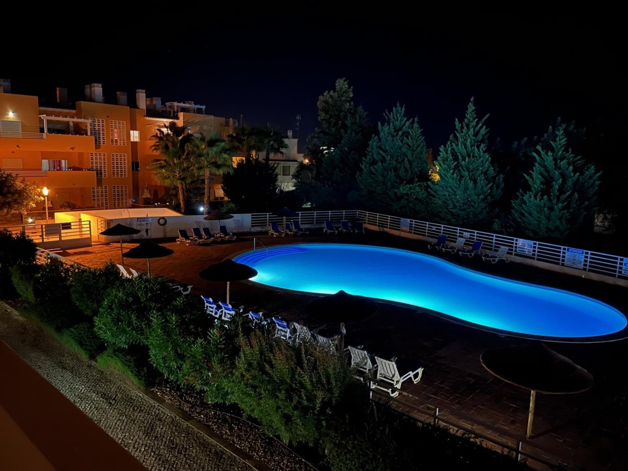 A nighttime swimming pool with lounge chairs and umbrellas, surrounded by trees and buildings.