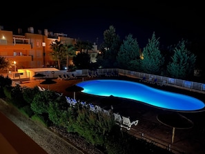 A nighttime swimming pool with lounge chairs and umbrellas, surrounded by trees and buildings. - Spacious apartment with air conditioning - 6 guests (Tavira)