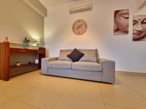 A living room with a sofa, a wall clock, a shelf with decorative items, and a wall-mounted air conditioner. - Spacious apartment with air conditioning - 6 guests (Tavira)