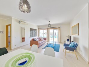 A modern living room with a sofa, a dining table, and a TV. There is a view of a pool through the glass doors. - Stylish apartment with air conditioning - 4 guests (Tavira)
