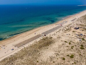 Oversigtsbillede af en strand med rækker af liggestole og parasoller, en sandstrand og havet i baggrunden.