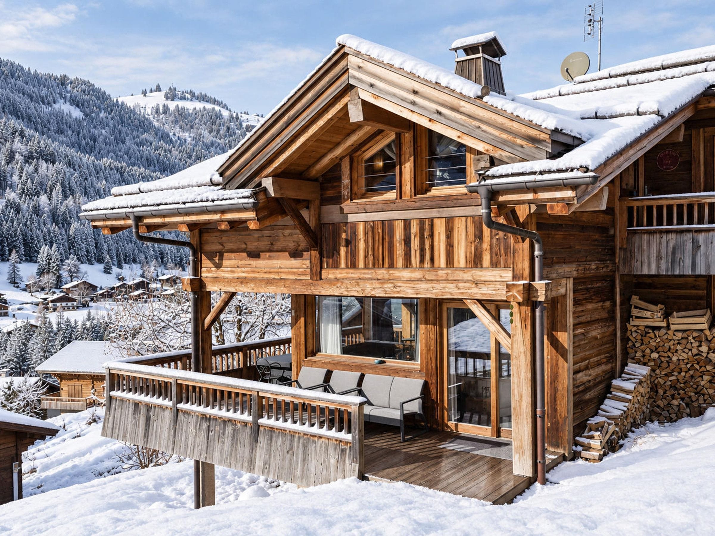 A wooden chalet with a balcony, large windows, and a view of a snowy mountain landscape.