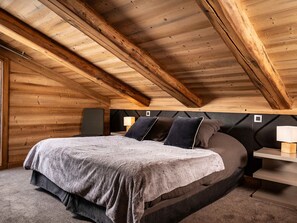 A bedroom with a sloped ceiling, wooden beams, a bed with a grey blanket, two bedside tables with lamps, and a dark headboard. - Chalet pets welcome - 8 guests, mountain view (La Clusaz)