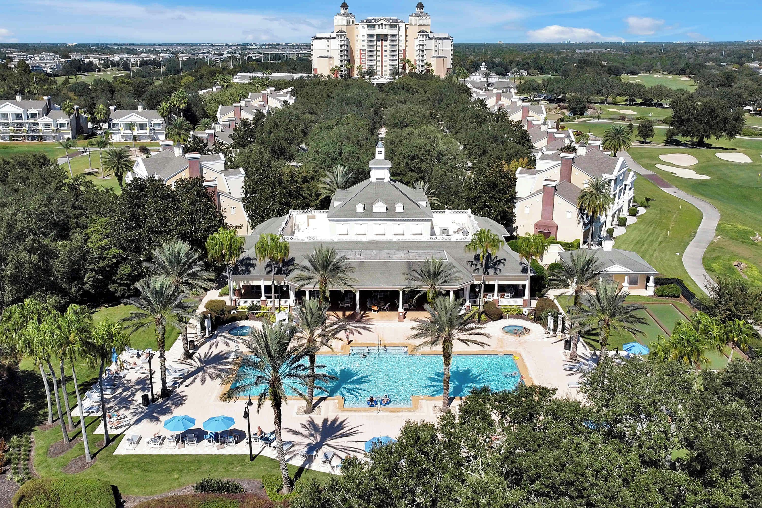 A large resort with a central pool, surrounded by palm trees and a golf course in the background.