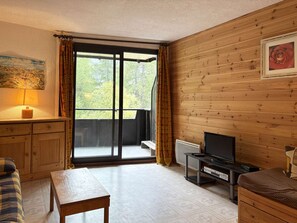 A room with a wooden interior, a television on a stand, a bench, a sofa, and a window with curtains. - Cozy studio with forest view - 4 guests (Les Orres)