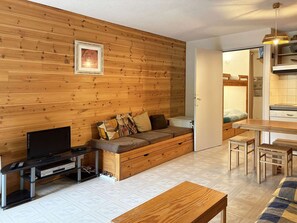 A cozy room with a wooden wall, a sofa with cushions, a TV, a wooden table, and a bench. - Cozy studio with forest view - 4 guests (Les Orres)