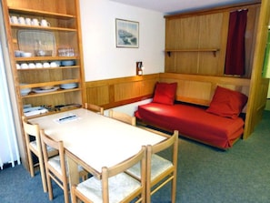 A compact room with a dining table, chairs, a built-in bench with cushions, a shelf with dishes, and a framed picture on the wall. - Sunny studio with balcony - 4 guests (Saint-Chaffrey)