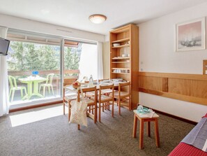 A room with a dining table set for four, a wooden bookshelf, a television on the wall, and a sliding glass door leading to an outdoor area with chairs. - Charming studio with South balcony - 4 guests (Saint-Chaffrey)