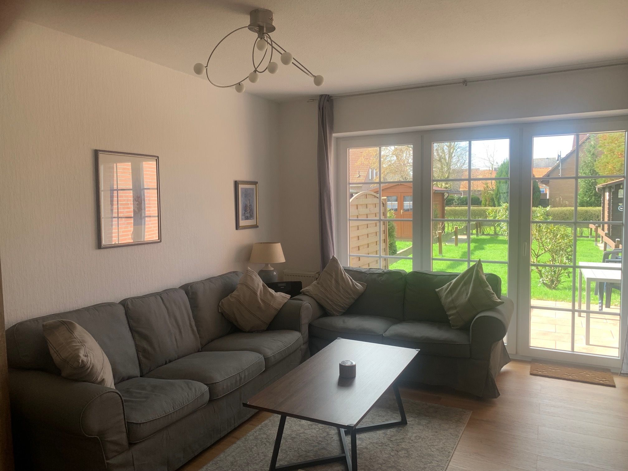 Ein Wohnzimmer mit grauem Sofa, einem Couchtisch und einer Lampe. Ein Fenster bietet Blick auf einen Garten und Häuser.