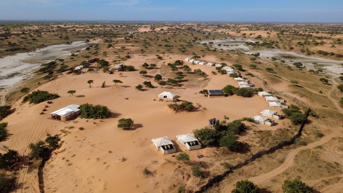 A circular arrangement of tents with a central building, surrounded by sparse vegetation and a sandy landscape.