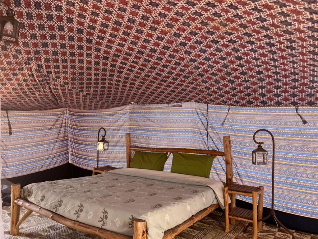 A tent with a patterned fabric interior, a wooden bed frame, and two wall-mounted lanterns.