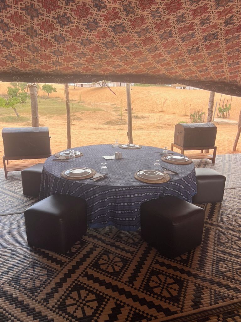 Une table ronde dressée pour le dîner, recouverte d’une nappe à motifs, entourée de chaises noires à coussins, et offrant une vue sur un espace extérieur avec des arbres et un bâtiment en arrière-plan.