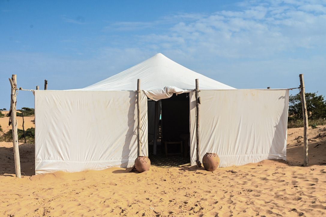 A white tent with a peaked roof, set on sandy ground with two large pots at the entrance.