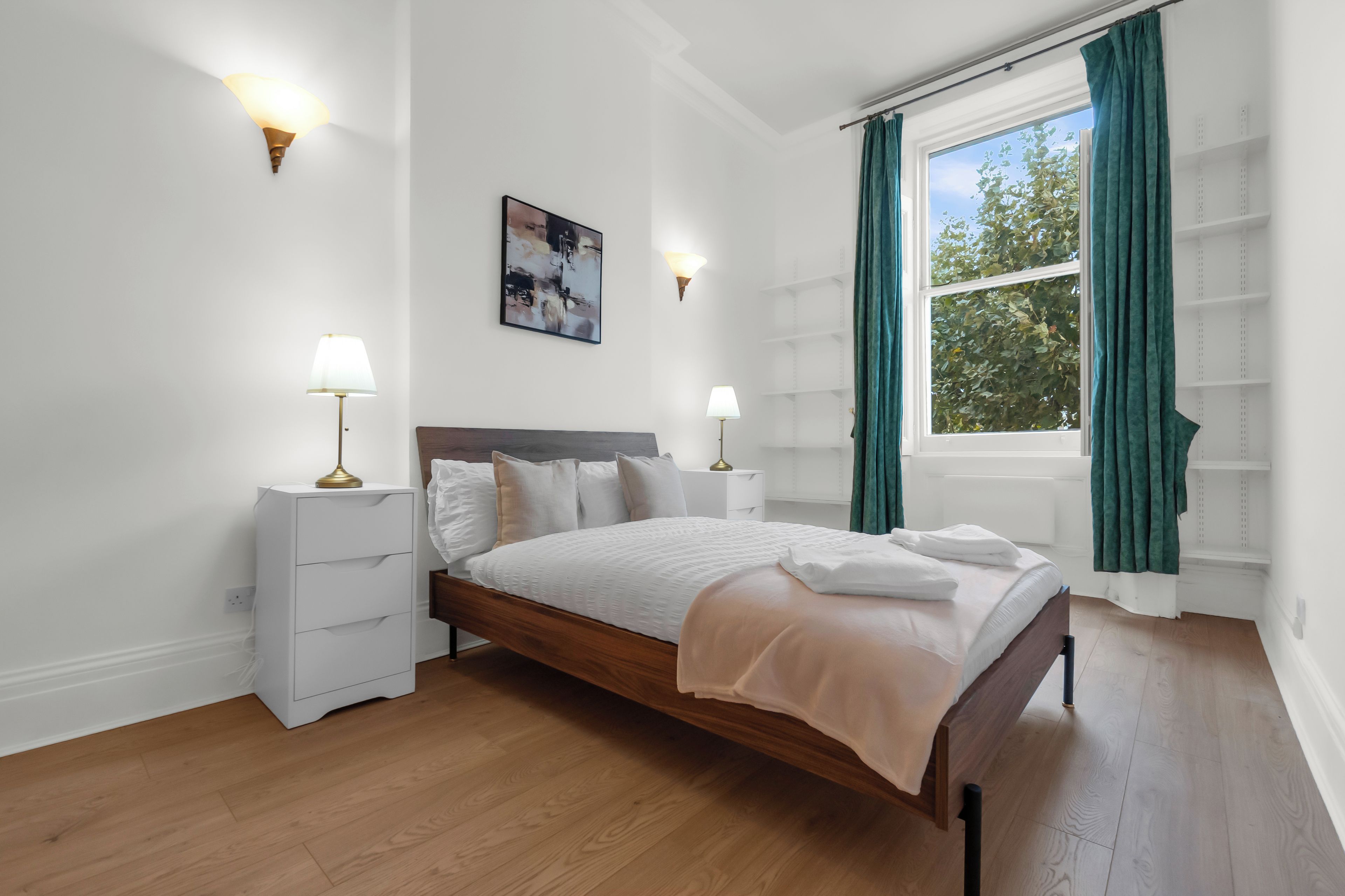Une chambre à coucher avec un lit en bois, des tables de chevet blanches, un tableau abstrait accroché au mur et une fenêtre avec des rideaux.