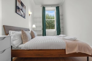A neatly made bed with pillows, a bedside table with a lamp, and a window with curtains. - Luxury South Kensington Retreat (London)