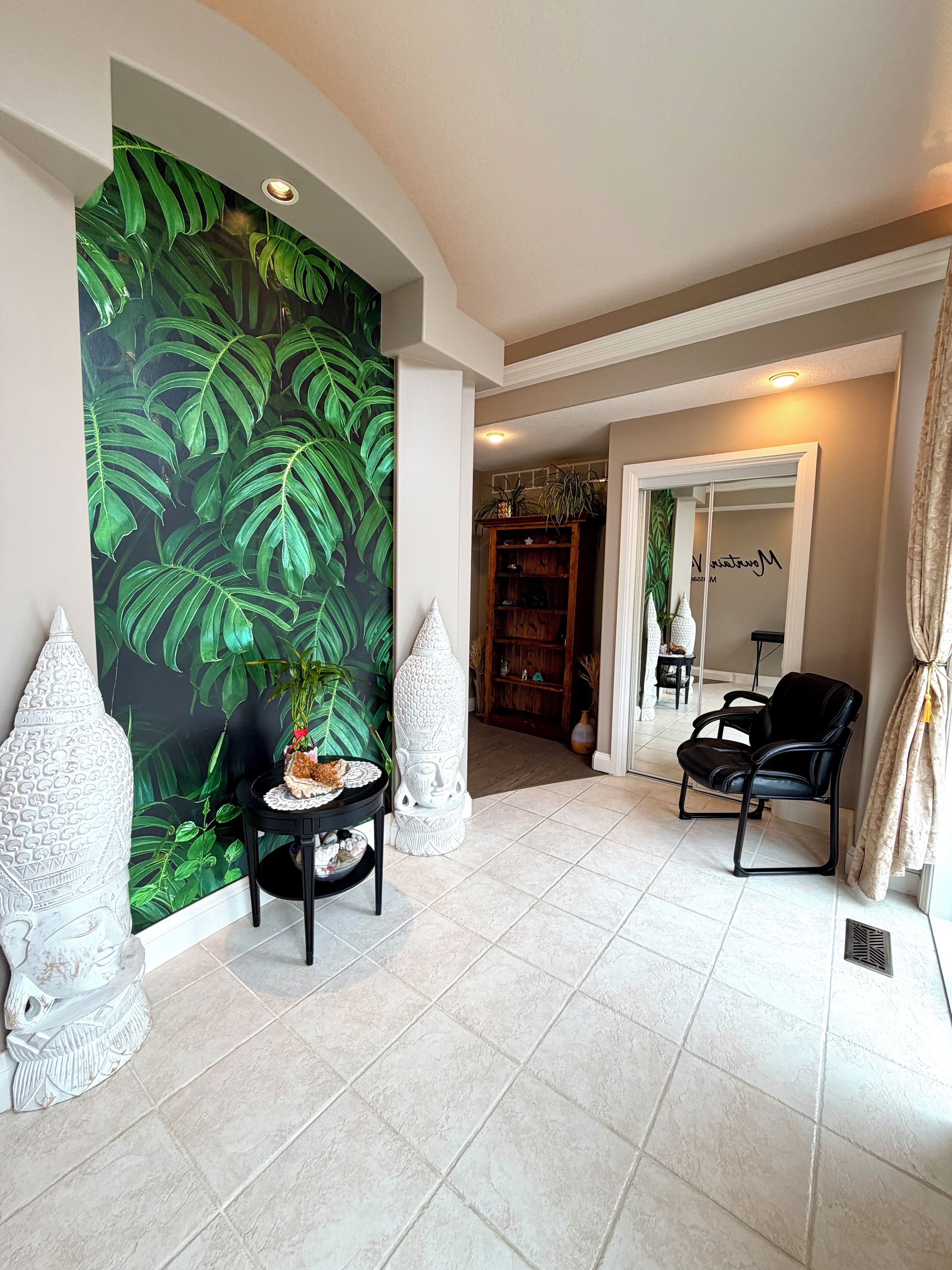 A room with a large green leafy wall mural, a black leather chair, a small round table with a decorative item, and a tall white vase.