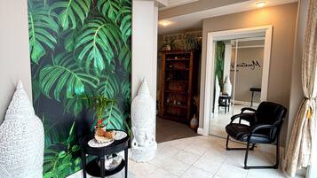 A room with a large green leafy wall mural, a black leather chair, a small round table with a decorative item, and a tall white vase.