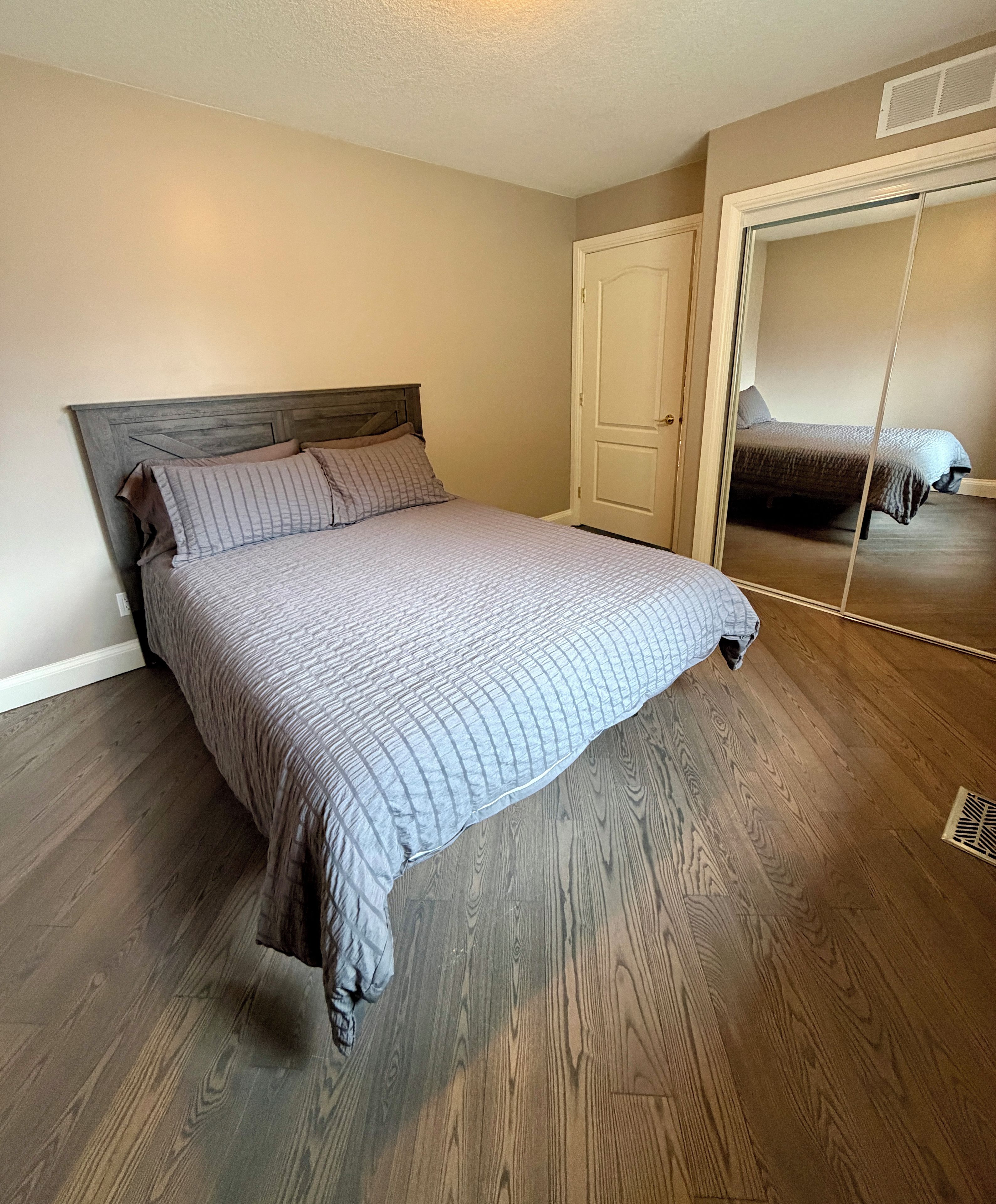 A bedroom with a bed, wooden flooring, a door, and a mirror.