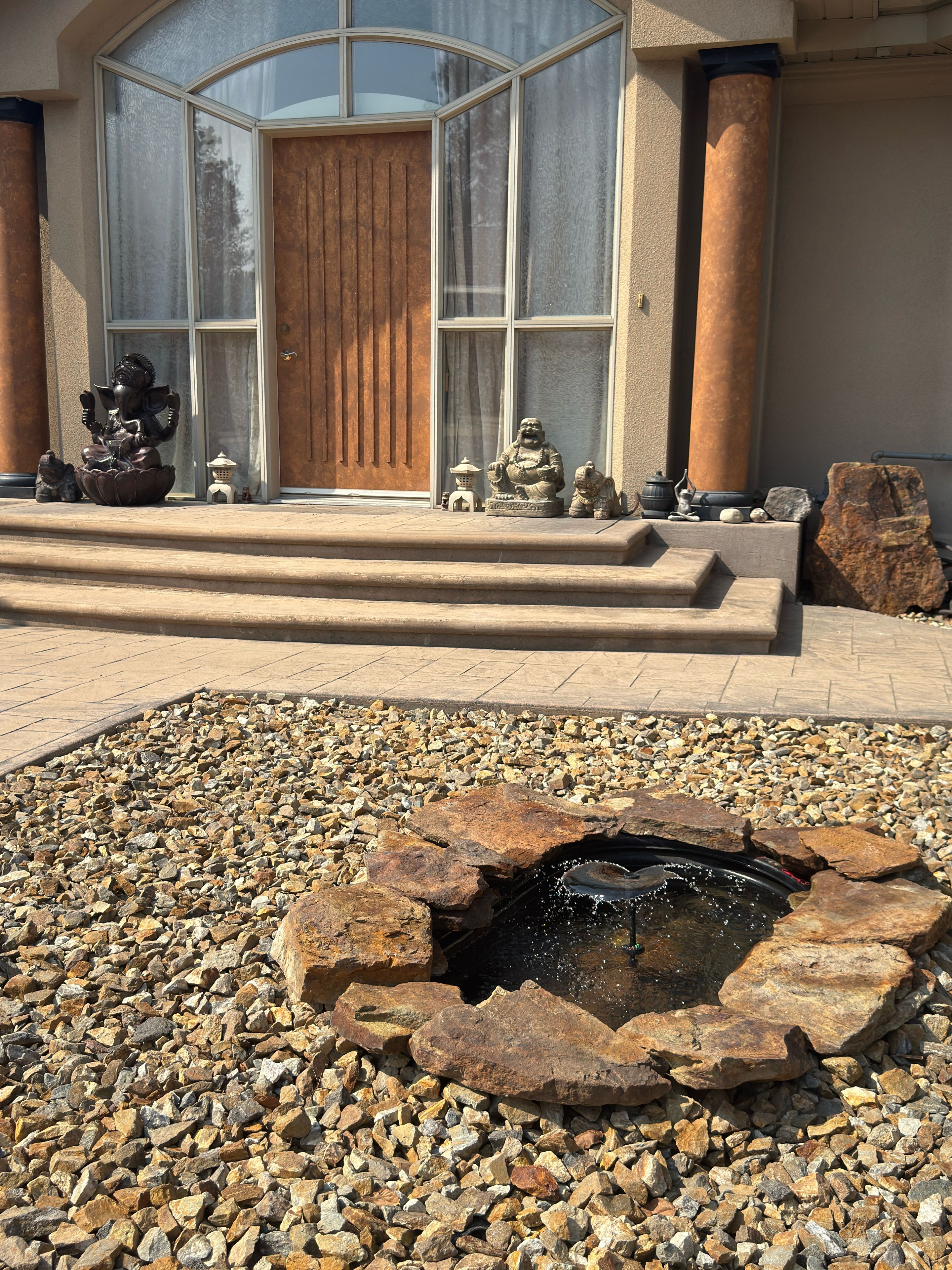A stone fountain with water, surrounded by pebbles, in front of a building with a wooden door and large windows.