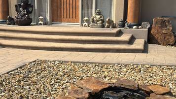 A stone fountain with water, surrounded by pebbles, in front of a building with a wooden door and large windows.