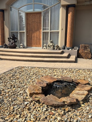 A stone fountain with water, surrounded by pebbles, in front of a building with a wooden door and large windows. - Spacious 3BR Knox Mountain Home | 5 Min to Downtown (Kelowna)