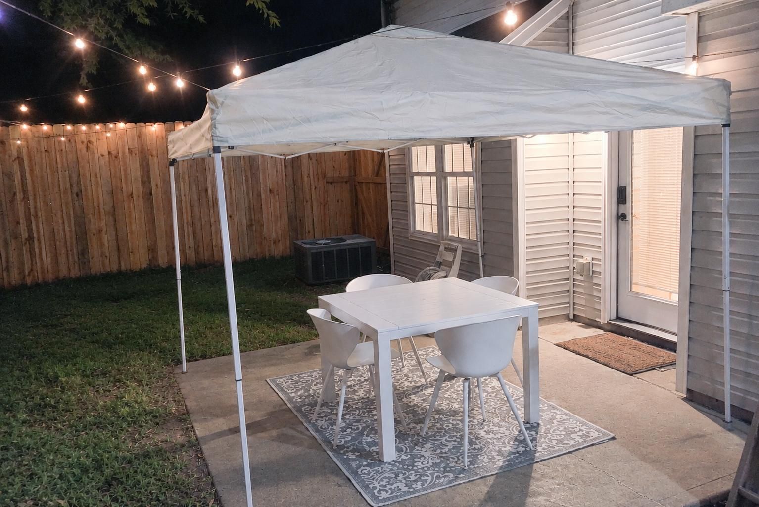 Een terras buiten met een witte tafel en stoelen, een overkapping en lichtslingers.