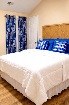 Een slaapkamer met een bed, witte beddengoed, blauw-witte gordijnen en een houten hoofdbord.