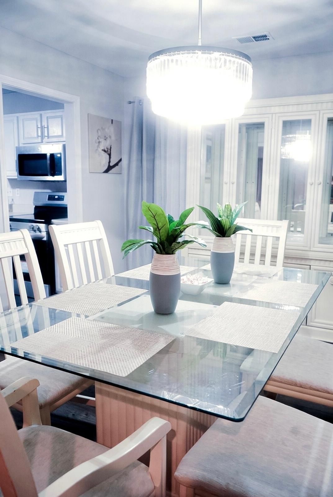 Un comedor con mesa de vidrio, sillas blancas y dos plantas en macetas.