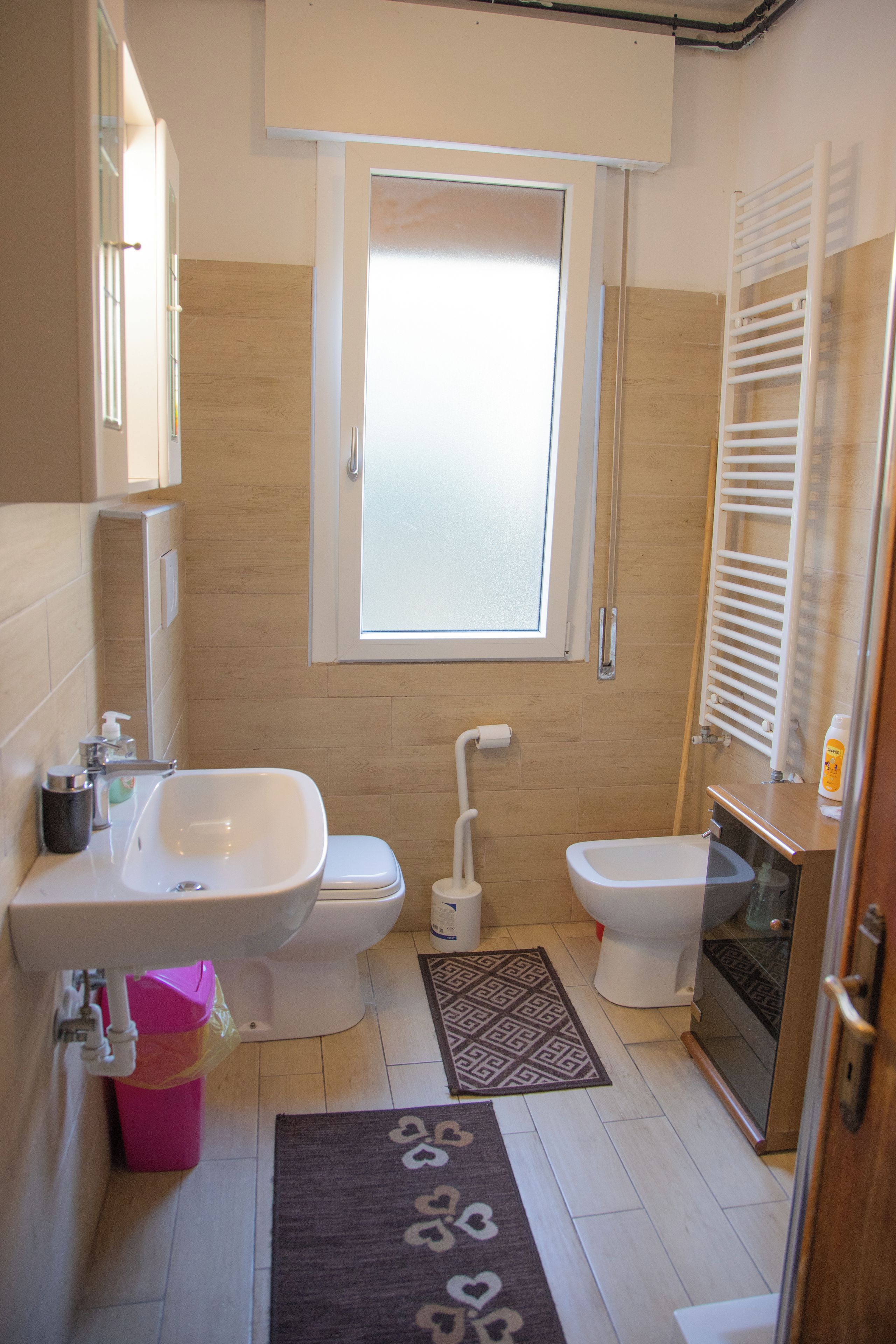 A compact bathroom with a sink, toilet, and bidet. A window provides natural light, and a patterned rug is on the floor.