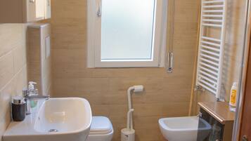 A compact bathroom with a sink, toilet, and bidet. A window provides natural light, and a patterned rug is on the floor.