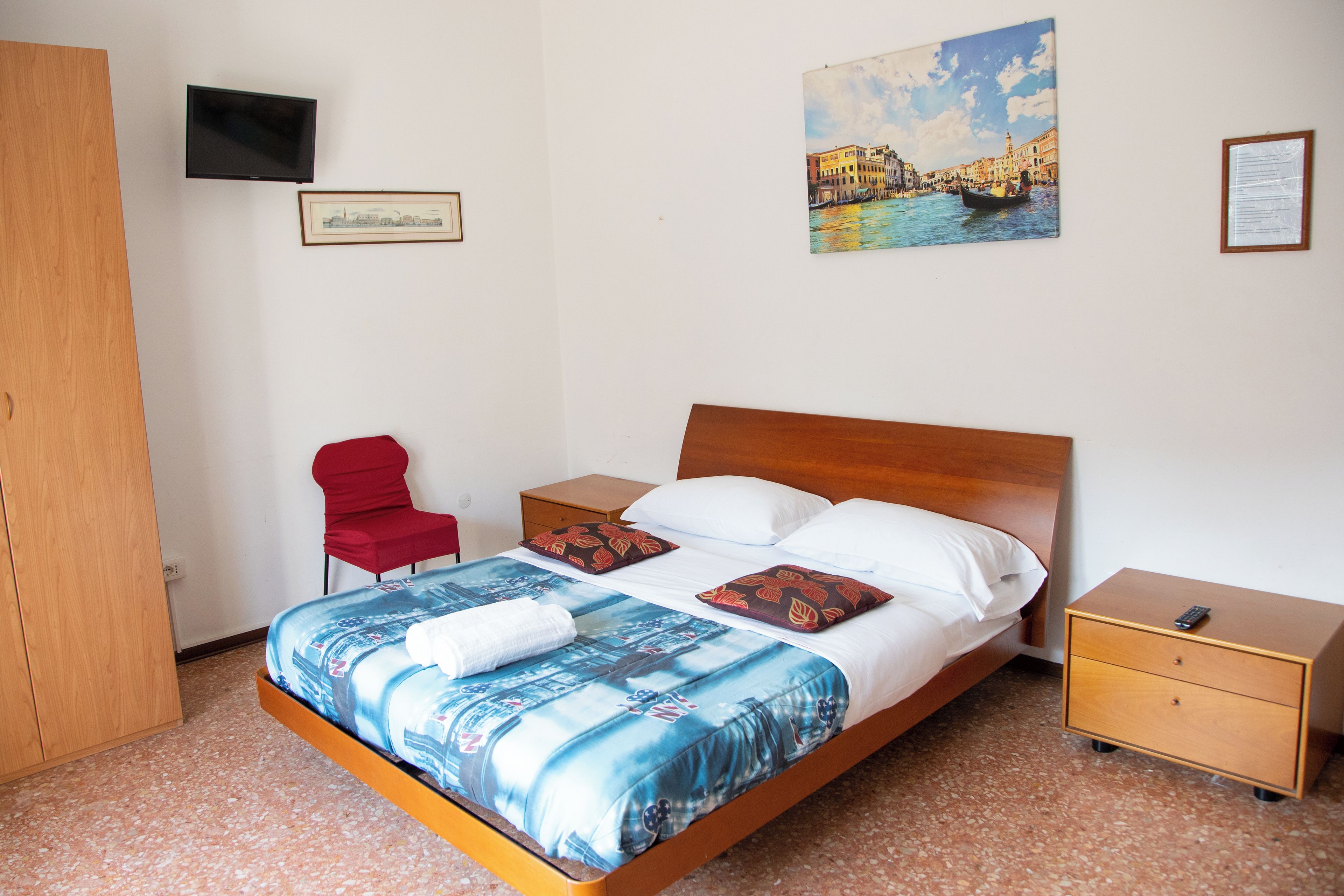 A hotel room with a bed, a TV, a red chair, a wooden wardrobe, and a painting of a cityscape.