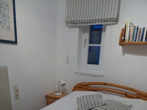 A bedroom with a wooden bed, a bedside table with a lamp, a window with a striped blind, and a bookshelf with books. - Holiday Apartment With 1 Bedroom (Wyk auf Foehr)