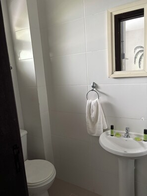 Un baño con inodoro, lavamanos y artículos de aseo, y una toalla colgada en un perchero.