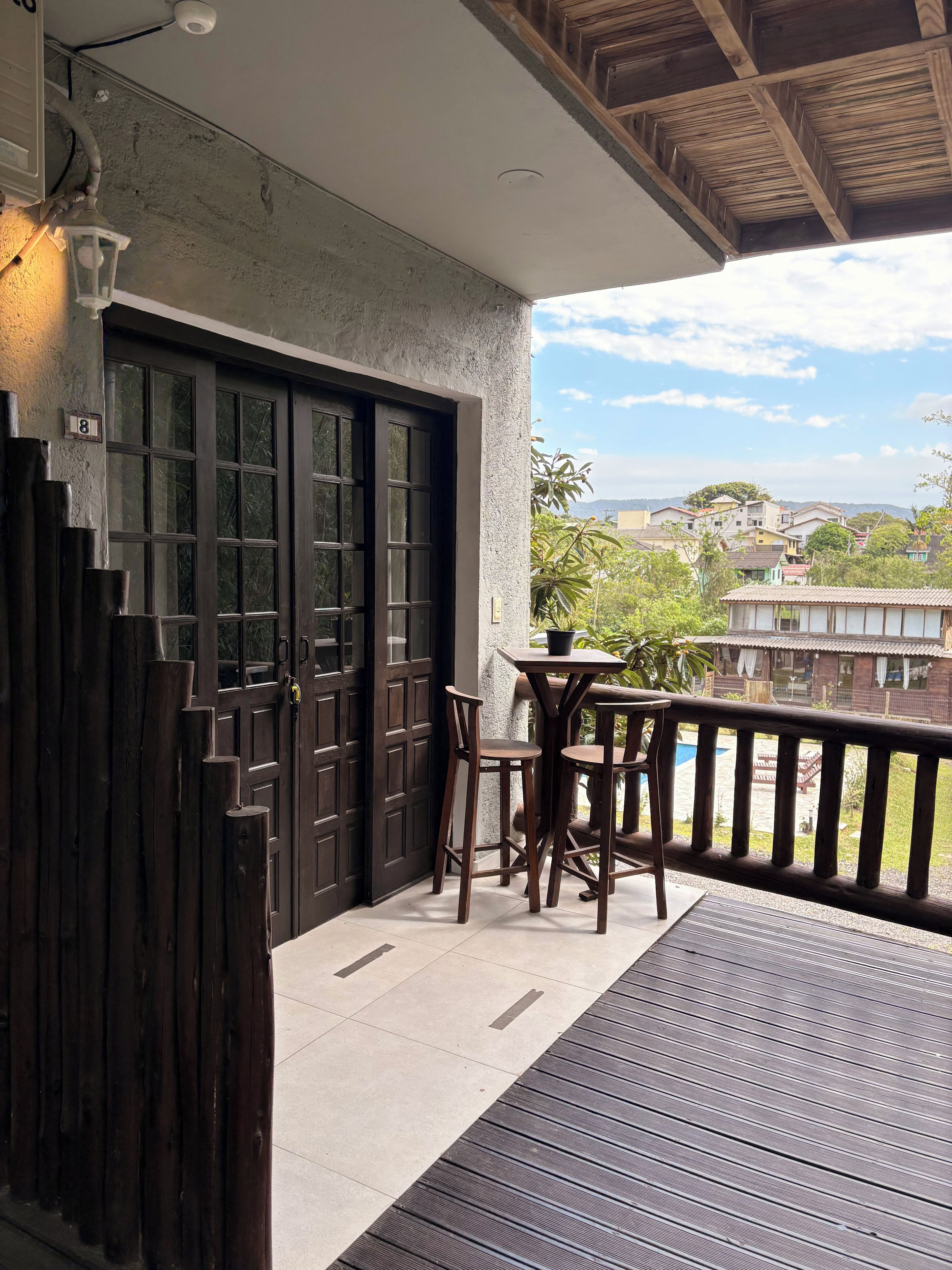 Un balcón de madera con una mesa y sillas, una puerta de madera oscura y vistas a una zona residencial.