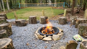 Un foyer entouré de pierres et de troncs d’arbres, avec une pelouse et un filet de volley-ball en arrière-plan.