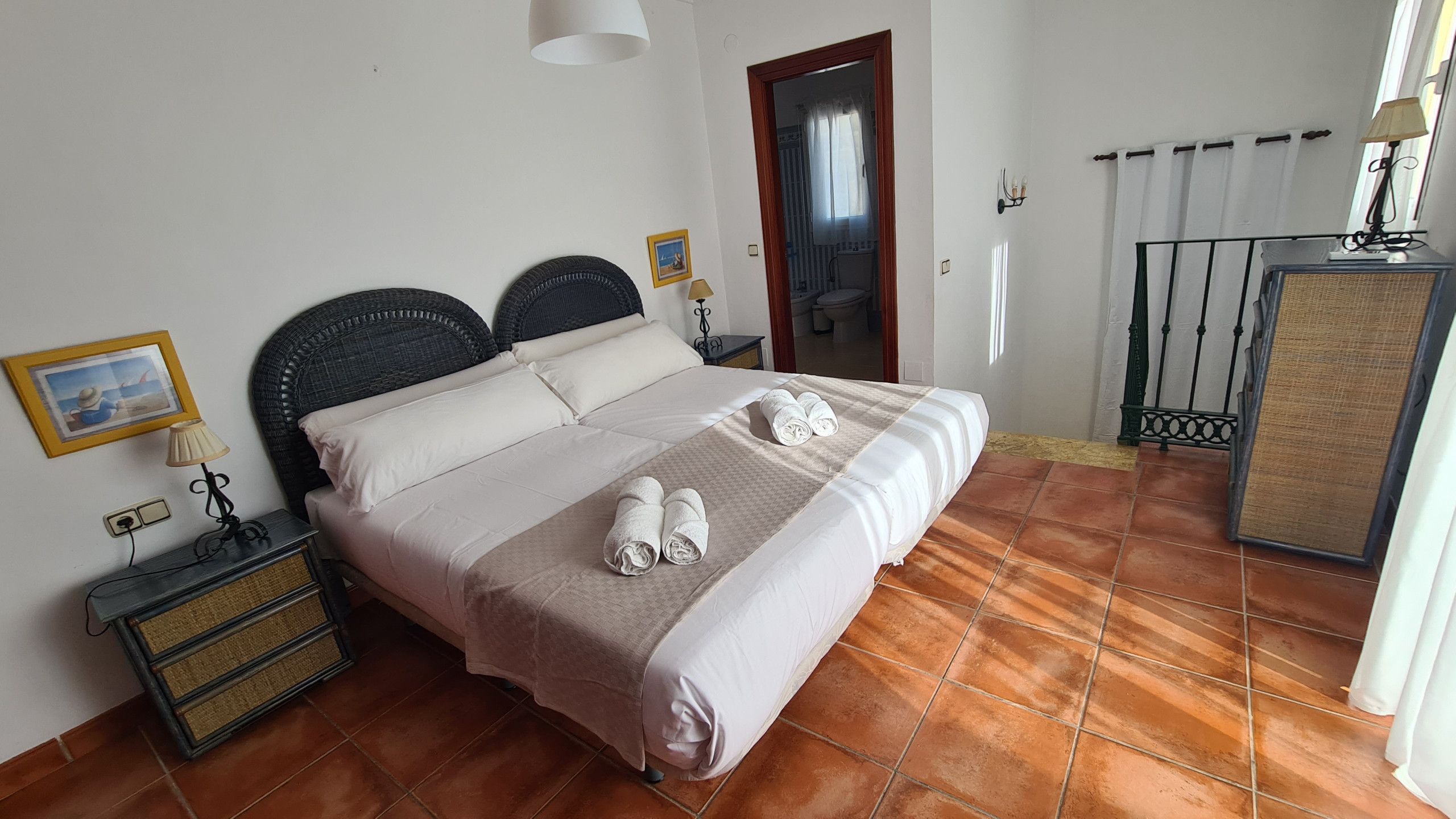 A neatly made bed with white linens and two rolled towels, a bedside table with a lamp, a framed picture on the wall, and a bathroom visible through an open door.