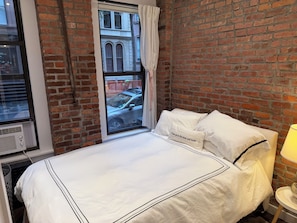 Een hotelkamer met een bakstenen muur, een groot raam met gordijnen, een bed met wit beddengoed en een nachtkastje met een lamp.