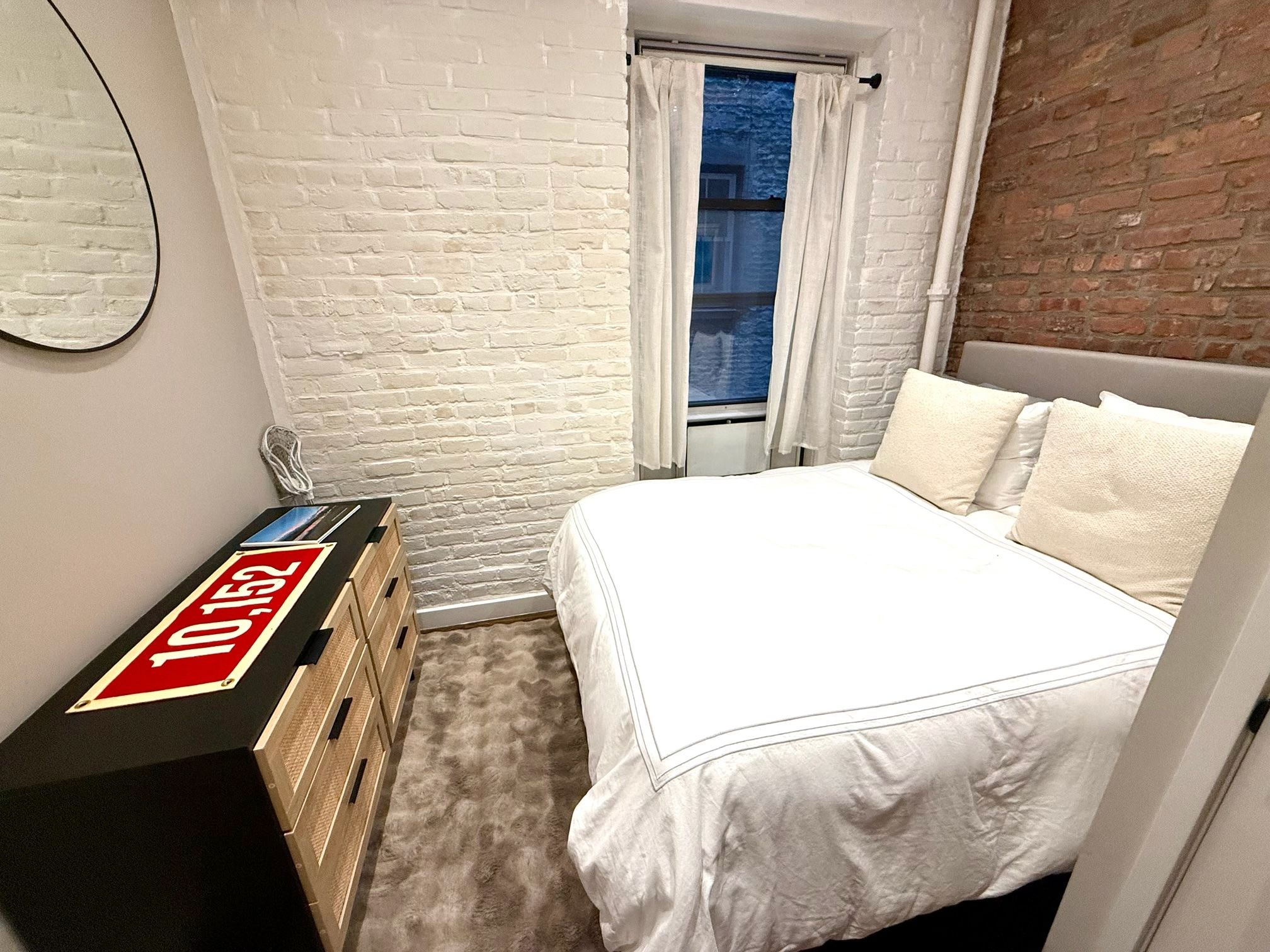A bedroom with a white brick wall, a bed with white bedding, a dresser with a red sign, and a window with curtains.