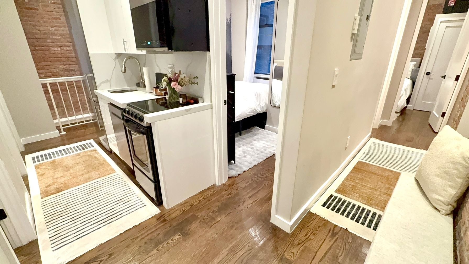 Cocina compacta con estufa, pileta y una pequeña mesada. Hay una cama con cubrecama blanco y una ventana con vista. El piso es de madera con alfombra estampada.