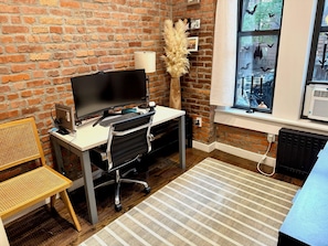 A home office with a brick wall, a desk with a computer, a chair, a lamp, and a window with a view of bats.