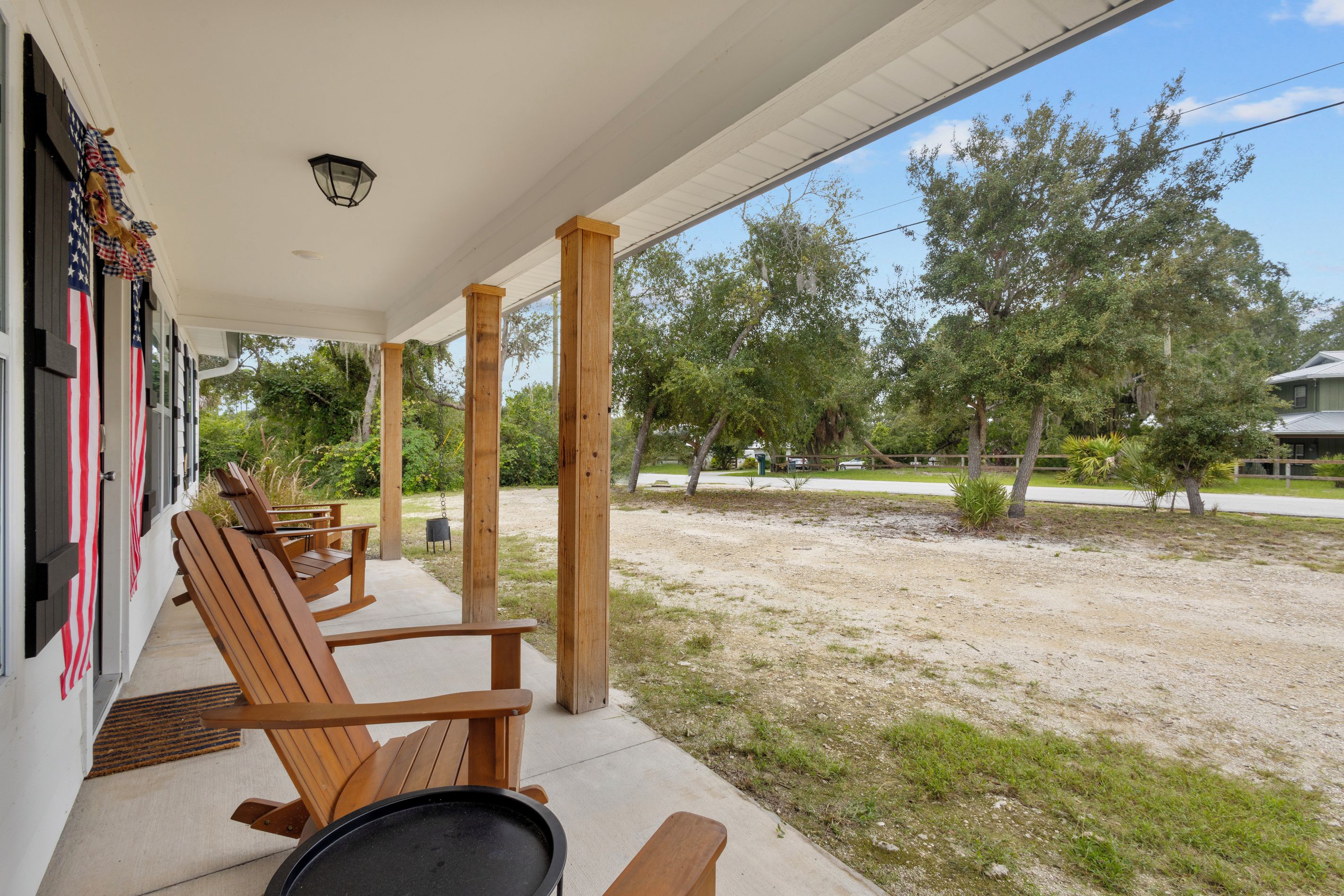 A covered outdoor seating area with wooden chairs and a table, overlooking a grassy yard with trees.