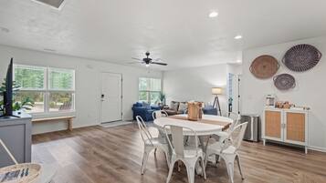 A spacious living area with a dining table and chairs, a ceiling fan, and a view of the outdoors through large windows.