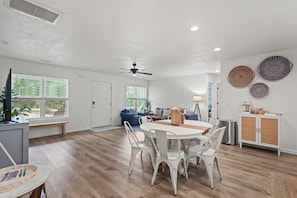 A spacious living area with a dining table and chairs, a ceiling fan, and a view of the outdoors through large windows.