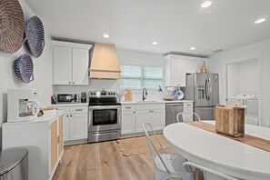 A modern kitchen with white cabinetry, stainless steel appliances, and a dining area with a wooden table. - Shelly's Chateau- Coastal Farmhouse w/ Fenced Yard (Cedar Key)