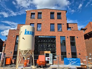 Een meerlaags gebouw van baksteen met een grote cilindrische tank waarop 'KNAUF' staat en een bouwplaats met hekken en materieel.